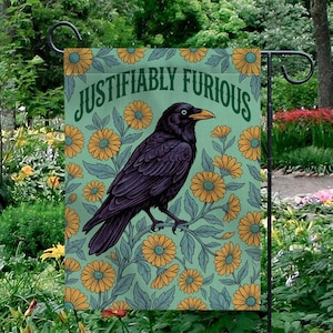 May include: A garden flag with a black raven design, set against a light green backdrop with yellow and orange floral patterns. The words "Justifiably Furious" are arched across the top. The flag is displayed on a black metal stand.