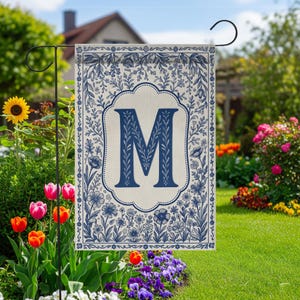 May include: A rectangular garden flag featuring a large, dark blue letter M centered within a decorative scalloped frame. The flag has a cream-colored background with a detailed border of blue botanical illustrations, including flowers and vines.