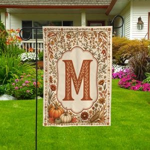 May include: A rectangular garden flag featuring a cream-colored background with a central scalloped frame containing a large, burnt orange letter M. The border is decorated with autumn-themed illustrations of pumpkins, corn, and foliage.