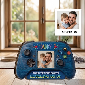 Op de afbeelding: Een blauwe fotolijst met houtstructuur in de vorm van een videogamecontroller met een foto van een kind en een volwassene. Boven de foto staat DADDY en onder de foto staat de tekst THANK YOU FOR ALWAYS LEVELING US UP in pixellettertype.