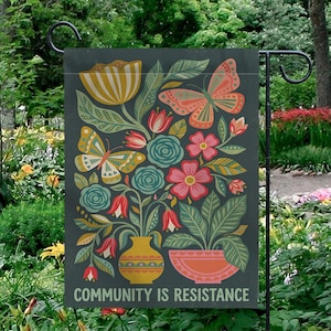 May include: A garden flag with a dark gray background features a floral design with butterflies, flowers, and foliage in shades of yellow, pink, and teal. The text "COMMUNITY IS RESISTANCE" is at the bottom.