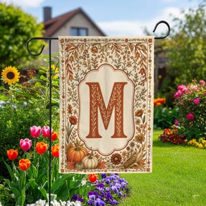 May include: A rectangular garden flag featuring a large, centered, rust-colored letter M inside a decorative scalloped frame. The border is adorned with autumn-themed illustrations including orange pumpkins, corn, and various fall-colored foliage.