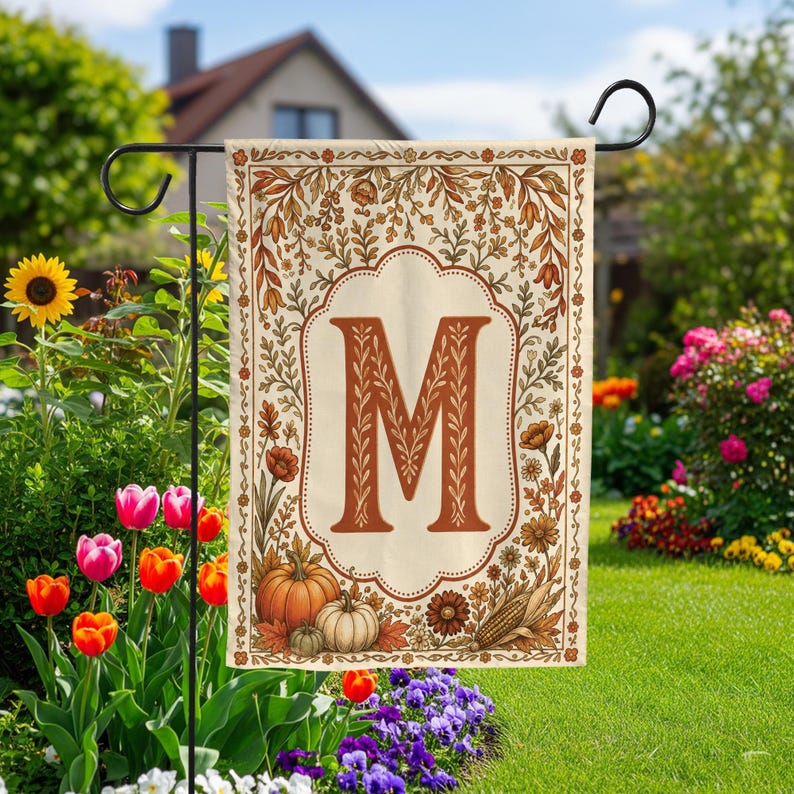 May include: A rectangular garden flag featuring a large, centered, rust-colored letter M inside a decorative scalloped frame. The border is adorned with autumn-themed illustrations including orange pumpkins, corn, and various fall-colored foliage.