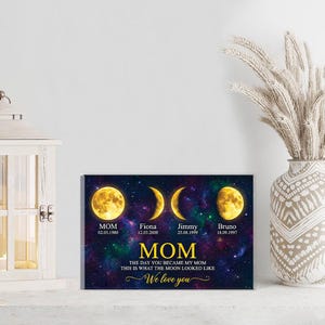 May include: A personalized canvas print featuring a celestial design with moon phases and names. The text reads "MOM" with dates and names below each moon phase. The phrase "The day you became my mom, this is what the moon looked like" is also included.