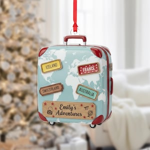 Puede incluir: Adorno navideño de maleta azul claro con diseño de mapa del mundo y detalles en rojo. El adorno tiene pegatinas de destinos de viaje para Islandia, Francia, Suiza y Australia. También se muestra el texto "Emily's Adventures".