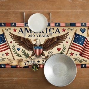 May include: A patriotic table runner with the word "AMERICA" and "250 YEARS" printed on it. The runner features an eagle, flags, stars, and hearts. Two white plates, a fork, and a knife are on the table runner.