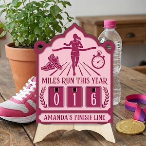 Puede incluir: Un letrero de hito de carrera de madera rosa y granate con el texto "MILES RUN THIS YEAR" y el número "016". El letrero presenta un gráfico de corredor, un cronómetro y un par de zapatillas para correr. También se ve una zapatilla para correr rosa, una botella de agua y una medalla.