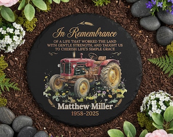 May include: A round, black memorial stone with the text "In Remembrance" and a red tractor illustration. The text includes a name and dates. The stone is surrounded by flowers and greenery.