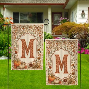 May include: Two rectangular garden flags featuring a cream background with a central scalloped frame containing a large orange letter M. The border is decorated with autumn-themed illustrations of pumpkins, corn, and various fall foliage in orange and brown.