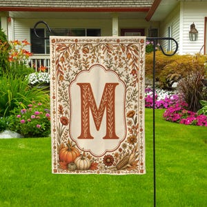 May include: A rectangular garden flag featuring a large, centered letter M in a serif font. The design is framed by a border of autumn-themed illustrations, including orange pumpkins, white gourds, corn, and various fall flowers and foliage.
