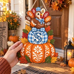 May include: A decorative hanging sign featuring a cartoon turkey wearing a blue gingham hat and bow tie, perched atop a blue gingham pumpkin with the letter S, resting on a large orange pumpkin with white floral patterns, surrounded by autumn leaves.