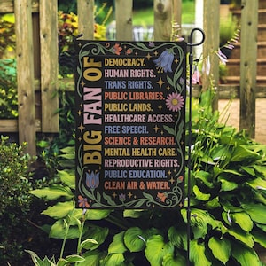 May include: A black garden flag with the words "BIG FAN OF" in gold lettering and a list of values in white. The flag is decorated with floral designs in various colors and is displayed in a garden setting.