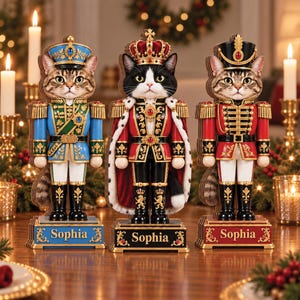 Può includere: Tre statuette schiaccianoci a forma di gatto su basi rettangolari. Il gatto a sinistra indossa un'uniforme blu e verde, quello centrale una veste reale rossa con corona e quello a destra un'uniforme rossa. Ogni base riporta il nome Sophia.