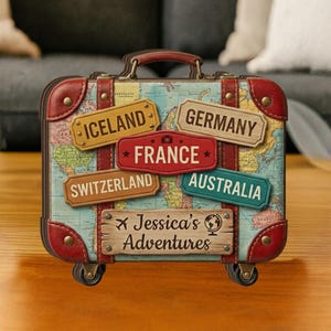 Może przedstawiać: Dekoracyjna walizka z mapą świata i etykietami miejsc docelowych. Walizka ozdobiona jest etykietami dla Islandii, Niemiec, Francji, Szwajcarii i Australii. Wyświetlany jest również tekst "Jessica's Adventures".