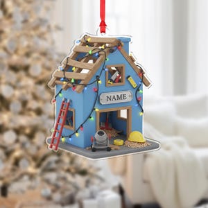 May include: A blue house-shaped Christmas ornament with a construction theme. It features a red ladder, wooden roof beams, and colourful string lights. The ornament has a sign that says "NAME".