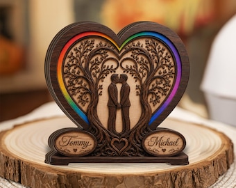 May include: A heart-shaped wooden ornament featuring two figures standing under intertwined trees. The design includes a rainbow-colored border and two oval plaques at the base engraved with the names Tommy and Michael in script font.