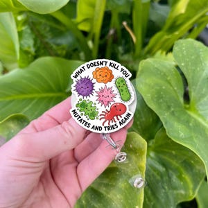 Puede incluir: Un carrete de insignia retráctil blanco con un diseño de dibujos animados colorido de varios microbios. El texto dice: "Lo que no te mata, muta y lo vuelve a intentar."
