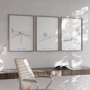 May include: Three framed art prints with minimalist line drawings. The prints feature landscapes with the words "THE WAY", "THE TRUTH", and "THE LIFE" at the bottom. The frames are a light brown color. A desk and chair are in the foreground.