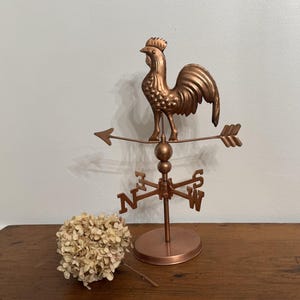 May include: A copper-colored metal weather vane featuring a rooster figure mounted on a horizontal arrow. Below the arrow, a directional cross displays the letters N, S, E, and W. The entire structure is supported by a circular base stand.