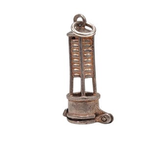 May include: A silver charm depicting a miner's lamp. The charm features a cylindrical base, a ladder-like structure, and a loop for attaching to a bracelet or necklace. The lamp is detailed with rivets and a small wheel.