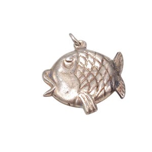 May include: A silver-colored fish charm with a textured, diamond-patterned body. The charm has a loop at the top for attaching to a necklace or bracelet. The fish has fins and an open mouth.