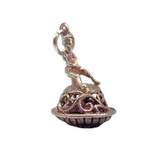 May include: A silver charm featuring a seated figure atop a decorative base with a purple gemstone. The base has an openwork design. The charm is suspended from a small loop, suitable for a necklace or bracelet.