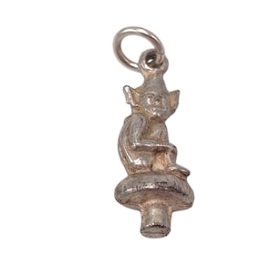 May include: A silver-toned charm depicting a seated gnome figure atop a mushroom. The gnome has pointed ears and is seated with its legs crossed. The charm has a loop for attachment and is likely a pendant or charm for a necklace or bracelet.