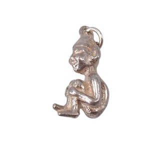 May include: A silver-colored charm in the shape of a seated figure with a pointed hat, holding its knees. The charm has a loop at the top for attachment to a necklace or bracelet. The figure is detailed with facial features and body contours.