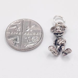 Pode incluir: Um pingente de metal prateado em forma de personagem de rato de desenho animado com orelhas grandes e bot&otilde;es redondos nos cal&ccedil;&otilde;es, posicionado ao lado de uma moeda brit&acirc;nica de cinco pence para escala. O pingente tem um pequeno anel.