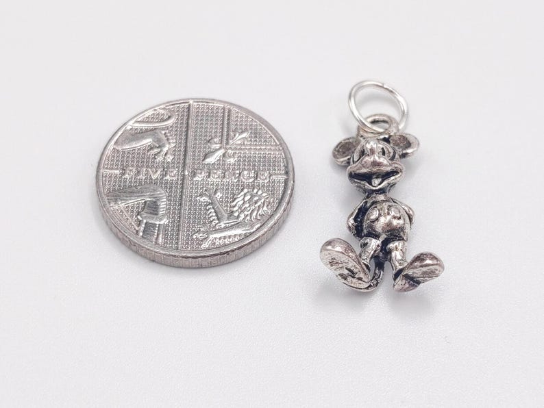Pode incluir: Um pingente de metal prateado em forma de personagem de rato de desenho animado com orelhas grandes e bot&otilde;es redondos nos cal&ccedil;&otilde;es, posicionado ao lado de uma moeda brit&acirc;nica de cinco pence para escala. O pingente tem um pequeno anel.