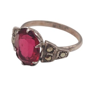 May include: Silver ring featuring a large, oval-cut, deep red gemstone. The ring has a detailed setting with smaller accent stones and decorative elements on the shoulders. The band is a simple, polished silver.