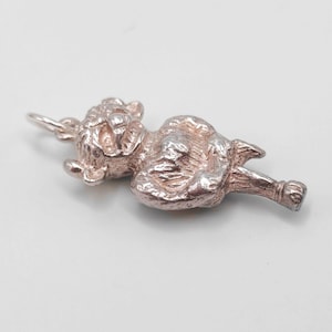 May include: A silver cat charm lying down, with detailed features. The charm has a small loop at the top for attaching to a chain or bracelet. The cat's face, paws, and tail are clearly visible. The charm is made of a silver-colored metal.