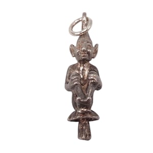Peut inclure: Un charm argenté représentant un gnome assis avec un visage souriant et des oreilles pointues, assis sur un champignon. Le charm a une petite boucle en haut pour l'attacher à un collier ou un bracelet.