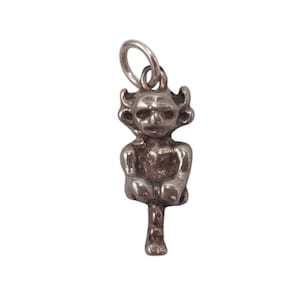 May include: A silver-toned pendant charm in the shape of a seated goblin or imp. The figure has a detailed face with horns and is suspended from a small loop. The charm is likely made of metal and has a vintage or antique appearance.