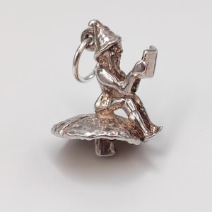 May include: A silver charm depicting a gnome reading a book while seated on a mushroom. The gnome wears a pointed hat and has a long beard. The mushroom has a textured cap and a smooth stem. The charm is designed to be attached to a bracelet or necklace.