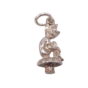 May include: A silver-colored charm depicting a seated figure atop a mushroom. The charm has a loop at the top for attachment to a necklace or bracelet. The figure is in a seated position with its arms wrapped around its knees.
