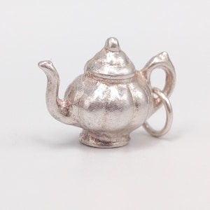 May include: A small, silver-colored teapot charm with a rounded body, curved spout, and a loop for attachment. The teapot has a lid and a handle. The charm is likely made of metal and is set against a white background.