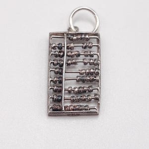 Puede incluir: Un dije de ábaco plateado con un pequeño lazo en la parte superior. El marco rectangular contiene múltiples filas de cuentas. Las cuentas son una mezcla de colores oscuros y claros. El dije es probablemente para un collar o pulsera.