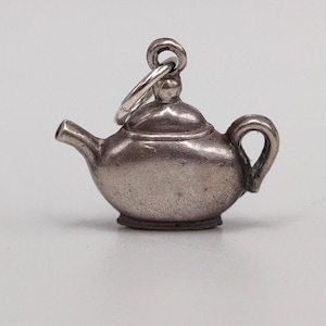 May include: A small, silver-toned teapot charm with a loop for attaching to a bracelet or necklace. The teapot has a spout, handle, and lid, and is likely made of sterling silver. The charm is a miniature version of a classic teapot.
