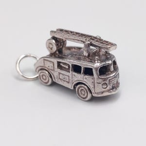 Op de afbeelding: Een zilverkleurige metalen bedel in de vorm van een vintage brandweerwagen. De bedel heeft een gedetailleerd uiterlijk met een ladder op het dak, ronde wielen en een kleine ring aan de achterkant om aan een armband of ketting te bevestigen.