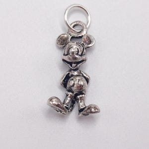 Pode incluir: Um pingente de metal em tom prateado com o formato do personagem Mickey Mouse, apresentando um design tridimensional detalhado com express&atilde;o sorridente, orelhas redondas e shorts. O pingente inclui um pequeno anel circular preso no topo.