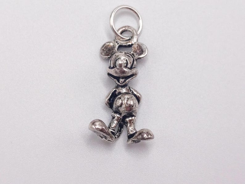 Pode incluir: Um pingente de metal em tom prateado com o formato do personagem Mickey Mouse, apresentando um design tridimensional detalhado com express&atilde;o sorridente, orelhas redondas e shorts. O pingente inclui um pequeno anel circular preso no topo.