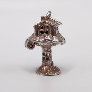 Könnte beinhalten: Ein silberfarbener Anhänger in Form eines skurrilen Hauses auf einem pilzartigen Sockel. Das Haus hat einen Schornstein und Fenster, und der Anhänger hat eine Schlaufe zum Befestigen. Der Anhänger ist detailliert und hat ein gealtertes Aussehen.