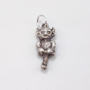 May include: A small, silver-coloured charm in the shape of a seated goblin or imp. The charm has a detailed face with horns and a mischievous expression. It is attached to a small ring for hanging.