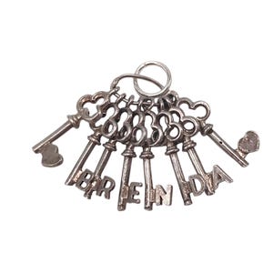 Könnte beinhalten: Eine Sammlung silberfarbener Schlüssel, jeder mit einem einzigartigen Design, verbunden durch einen Metallring. Die Schlüssel bilden den Namen "BRENDA". Die Schlüssel sind fächerförmig vor weißem Hintergrund angeordnet.