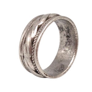Puede incluir: Un anillo plateado con un diseño texturizado. El anillo presenta un patrón trenzado en el centro, bordeado por una textura similar a una cuerda. El anillo tiene una apariencia ligeramente desgastada, lo que sugiere un estilo antiguo o vintage.