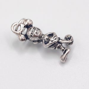Pode incluir: Um pingente de metal prateado representando um personagem de desenho animado com orelhas arredondadas, bra&ccedil;os cruzados e um pequeno anel no topo. A parte de tr&aacute;s da cabe&ccedil;a apresenta o texto "DISNEY" e "925" gravado no metal.
