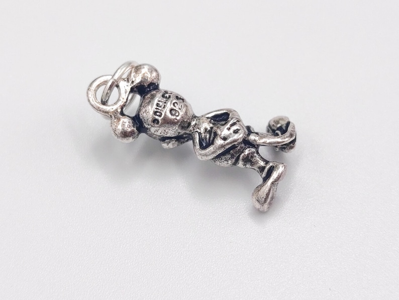Pode incluir: Um pingente de metal prateado representando um personagem de desenho animado com orelhas arredondadas, bra&ccedil;os cruzados e um pequeno anel no topo. A parte de tr&aacute;s da cabe&ccedil;a apresenta o texto "DISNEY" e "925" gravado no metal.
