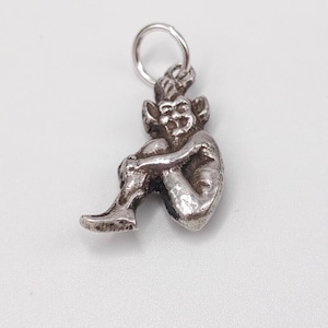 Puede incluir: Un pequeño dije plateado con forma de duende o goblin sentado. La figura está detallada con una cara sonriente, orejas puntiagudas y abraza sus rodillas. Un pequeño lazo en la parte superior para sujetarlo a un collar o pulsera.