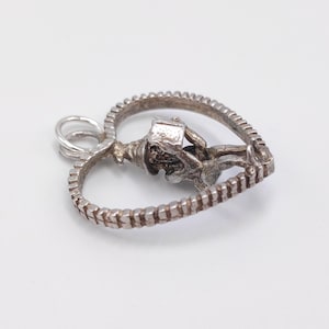 May include: A silver-toned heart-shaped pendant with a detailed design. The pendant features a textured border and a small figure holding a box. The bail is a simple loop for attaching to a chain.